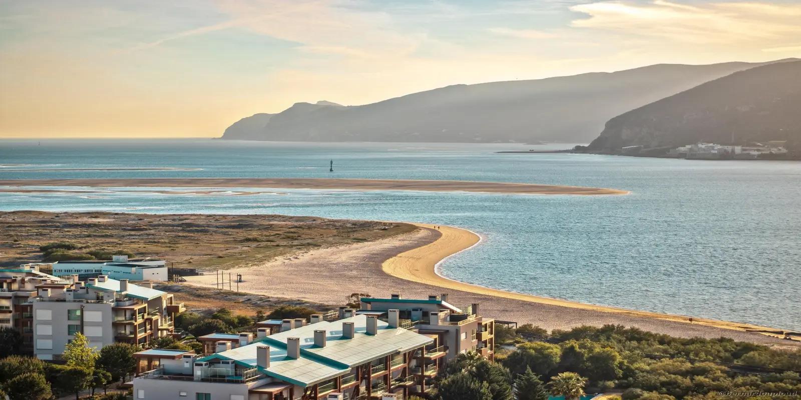 Scenic view of Setúbal, Portugal, featuring the Sado Estuary with a sandbar, calm blue water, and residential buildings in the foreground.