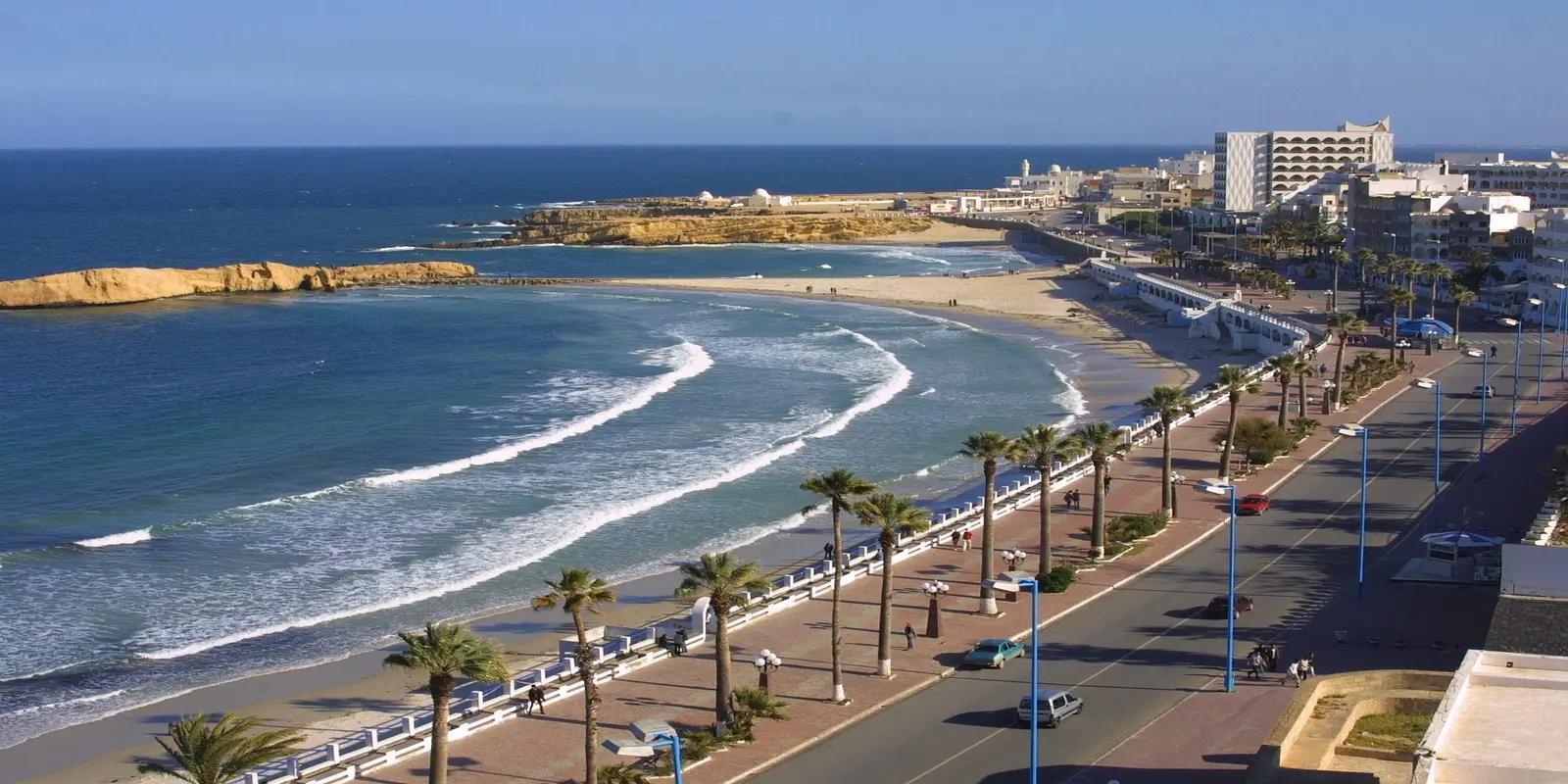 Aerial view of Al Qurayyah Beach in Saudi Arabia, featuring a sandy beach with gentle waves, a coastal road lined with palm trees, and the city skyline with buildings.