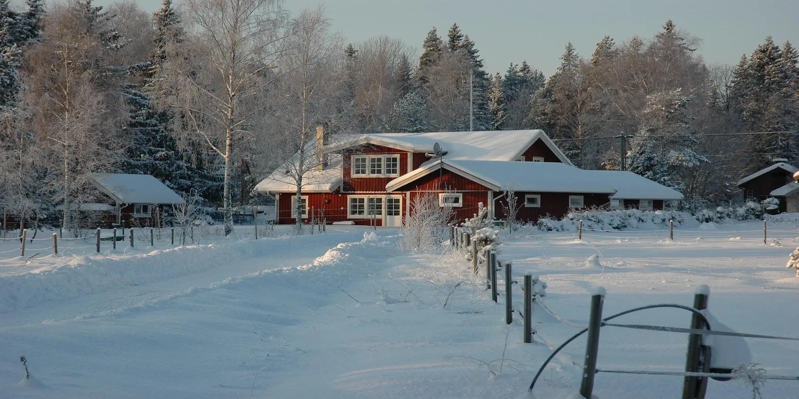A winter scene in Sweden featuring a traditional red wooden house with white trim, covered in snow, surrounded by snow-covered trees and a snowy landscape.