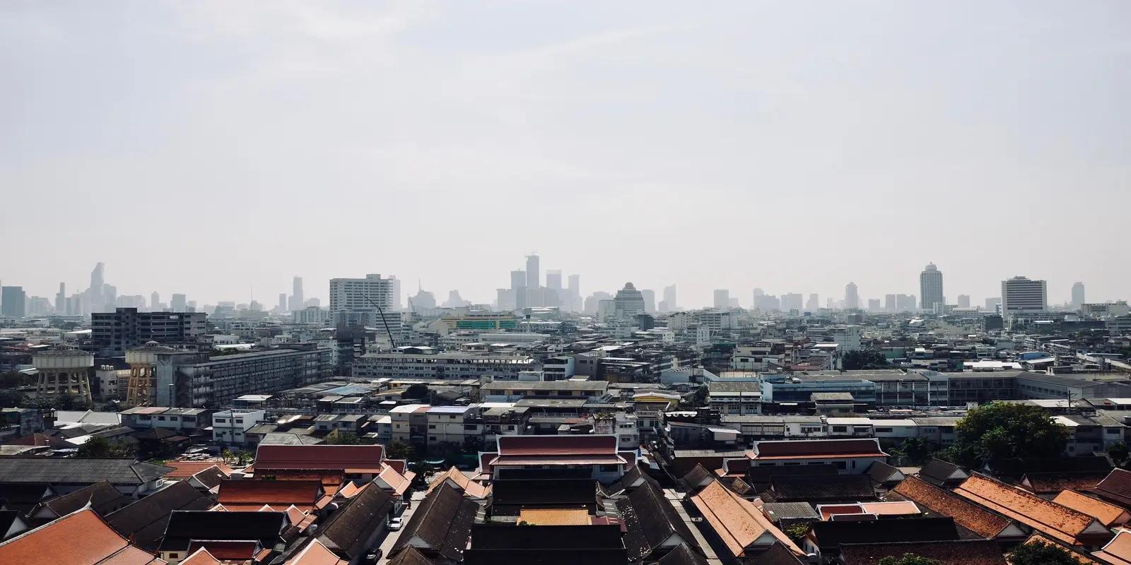 A panoramic view of Bangkok, Thailand, featuring dense urban development with traditional rooftops in the foreground and a hazy skyline with modern skyscrapers in the distance. The image captures the city's mix of old and new architecture under a bright, overcast sky.