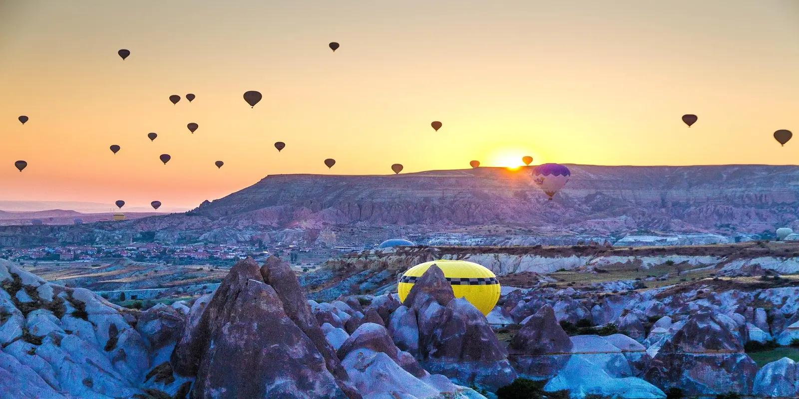 Sunrise view of Cappadocia, Turkey, with numerous hot air balloons silhouetted against a colorful sky. The landscape features unique rock formations and a valley, with the sun rising over the horizon. A yellow hot air balloon is prominently visible in the foreground.