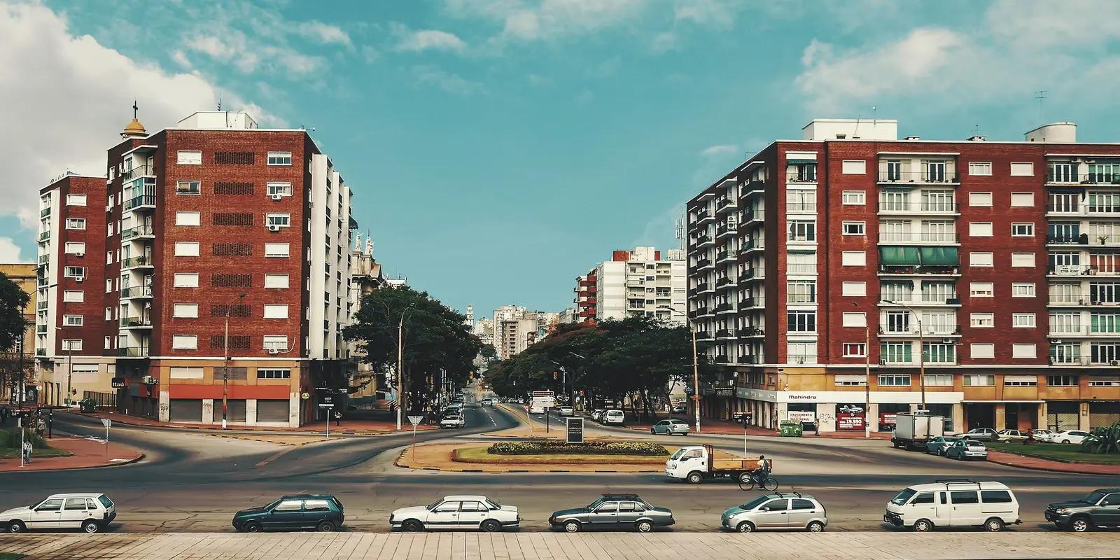 A sunlit urban street scene in Uruguay with two brick apartment buildings facing each other, creating a visual corridor. Cars are parked along the curb, and a small roundabout is visible in the middle of the intersection. The sky is blue with scattered white clouds.