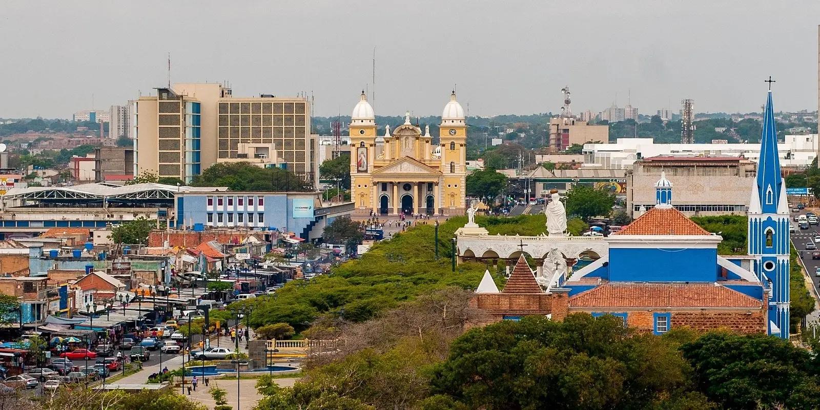 A cityscape of Maracaibo, Venezuela, featuring the yellow Basilica of Our Lady of Chiquinquirá, colorful buildings, busy streets, and a blue church tower in the foreground.