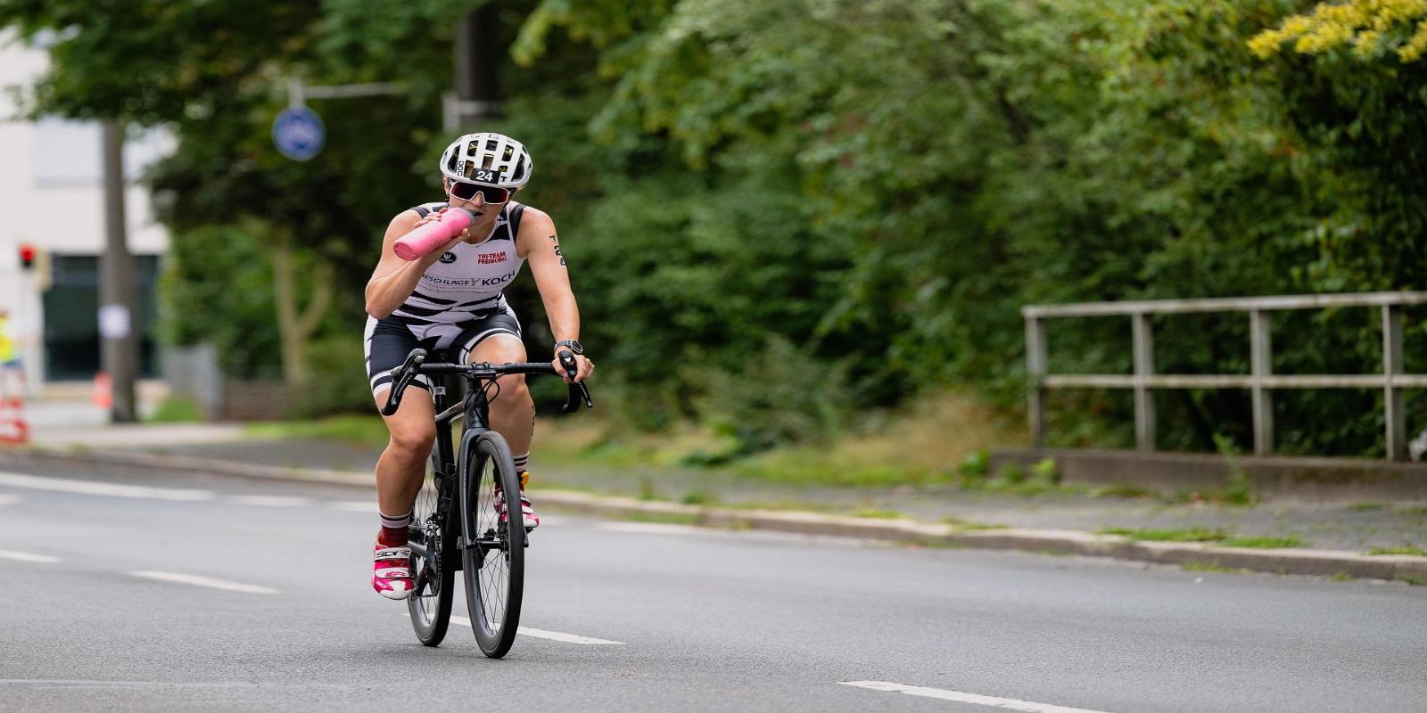 Determined woman cyclist in a triathlon race, wearing a helmet and sunglasses, drinking from a water bottle on a paved road with greenery.