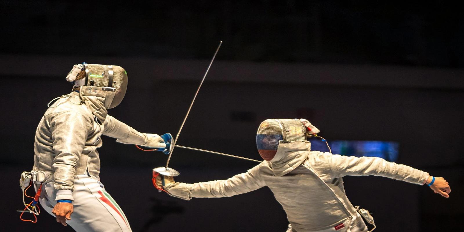 Two fencers in protective gear and masks engaged in a bout, swords crossed in a dynamic action shot on a dark stage.