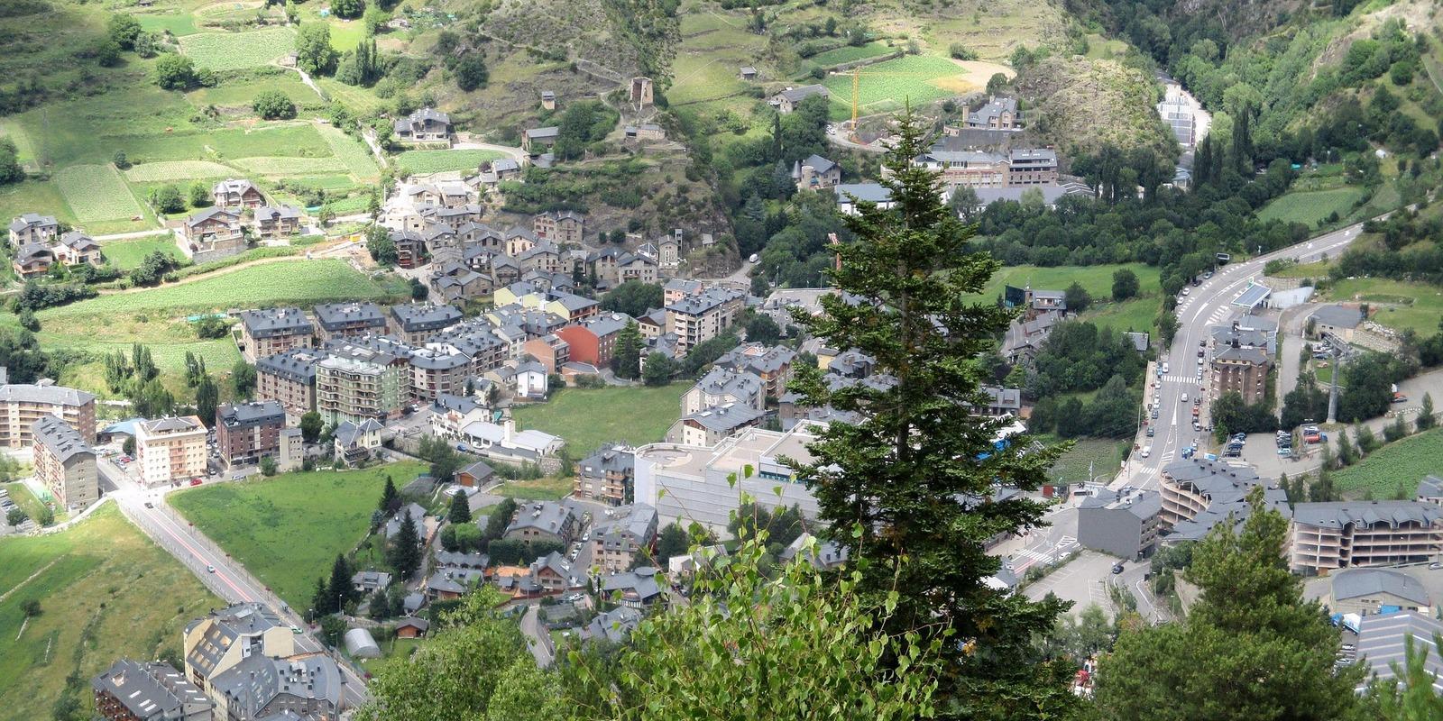 Panoramic view of a town nestled in a mountainous valley in Andorra, with traditional buildings, green fields, and winding roads under a clear sky.