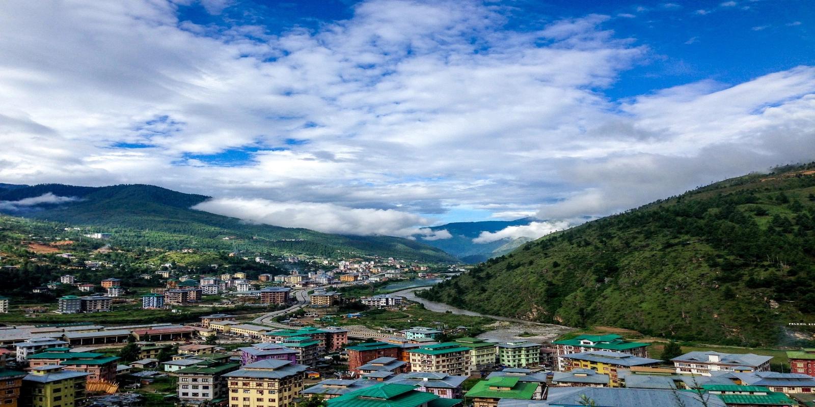 Aerial view of Thimphu city nestled in a lush green mountain valley in Bhutan, under a cloudy sky.