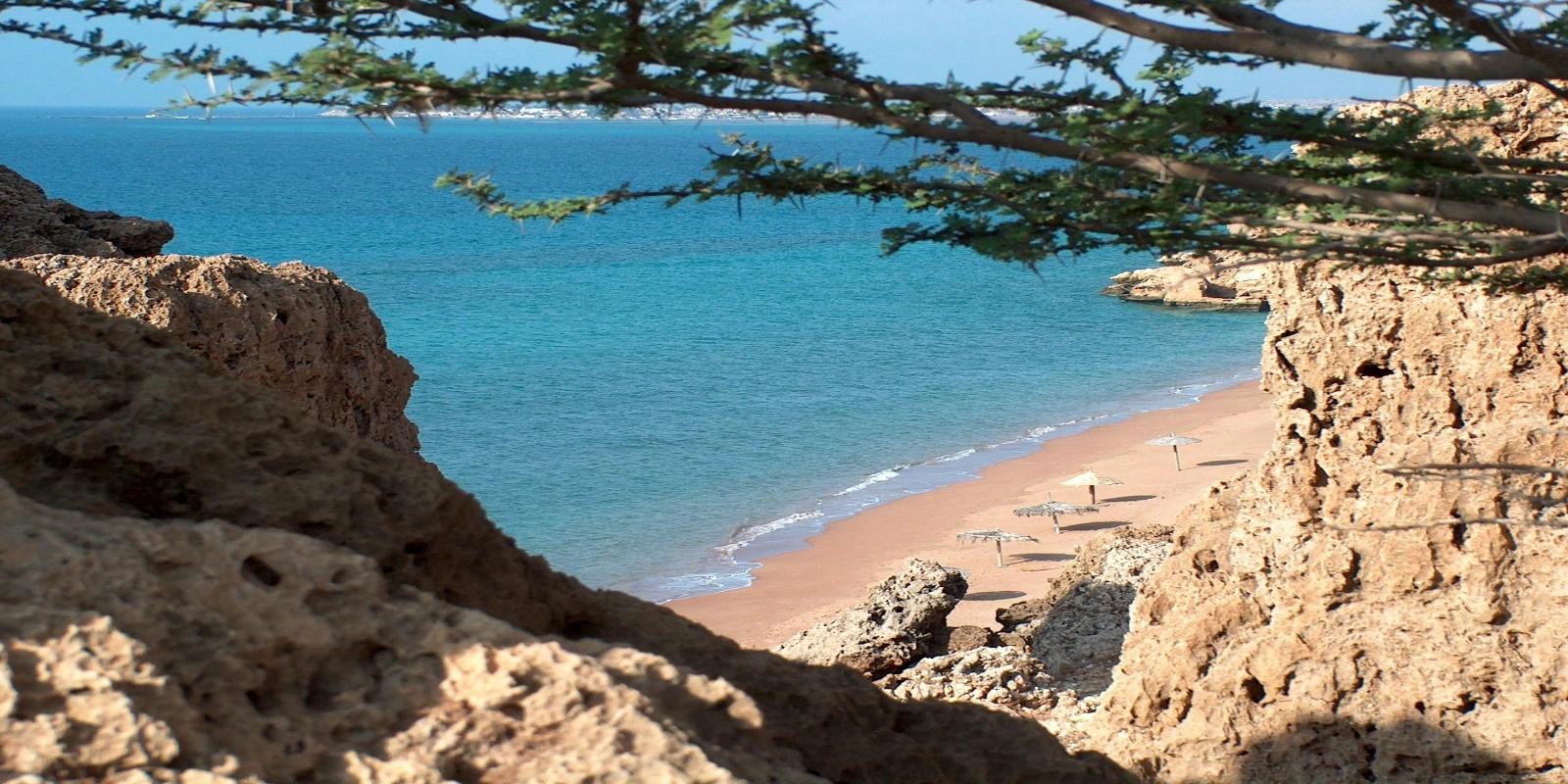 Secluded Ras Bir Beach in Djibouti, with calm turquoise waters and beach umbrellas, viewed from between rugged orange-brown cliffs.