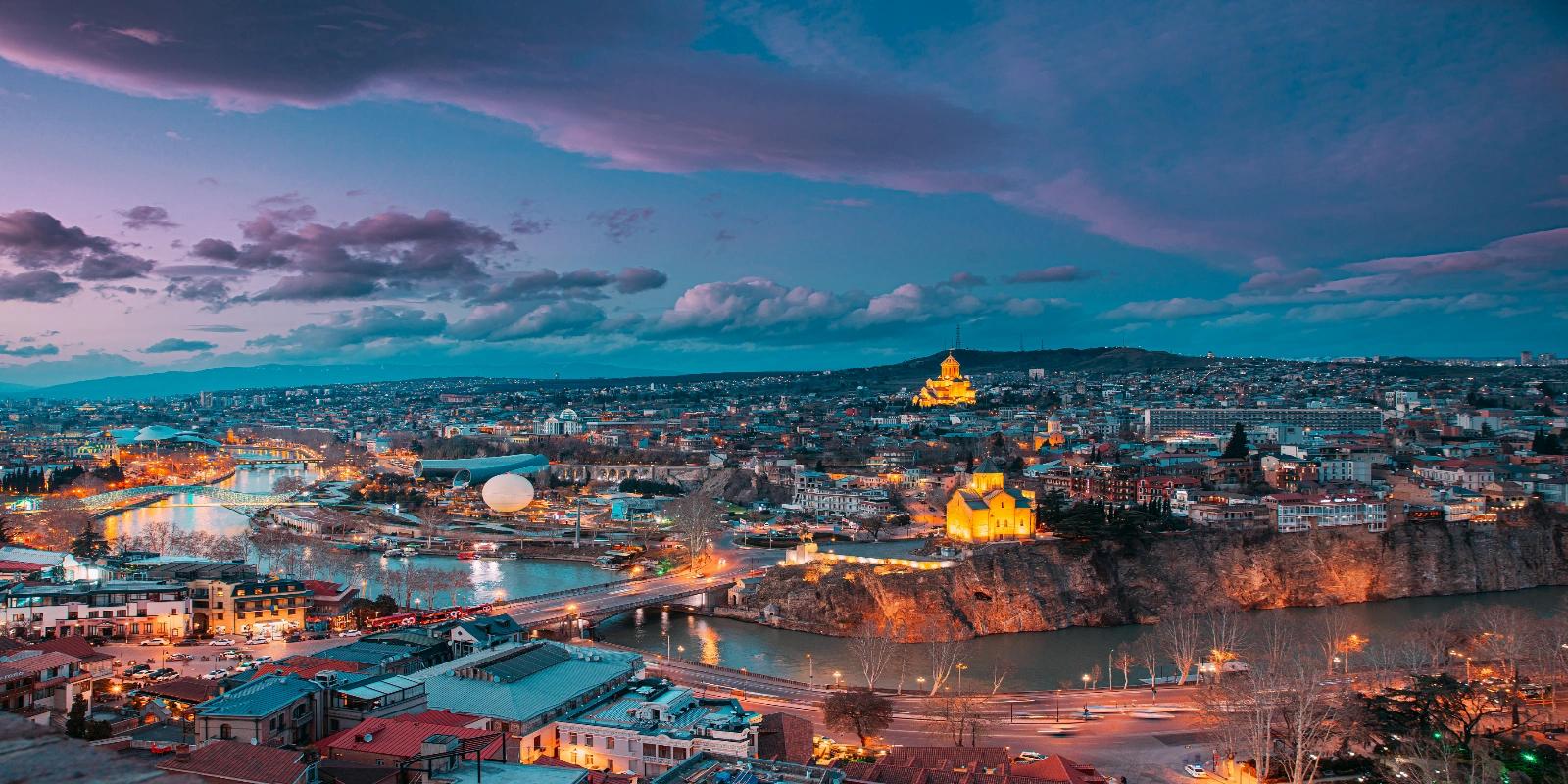 Enchanting twilight cityscape of Tbilisi, Georgia, with the Kura River, illuminated Narikala Fortress, and historic buildings under a dramatic, colorful sky.