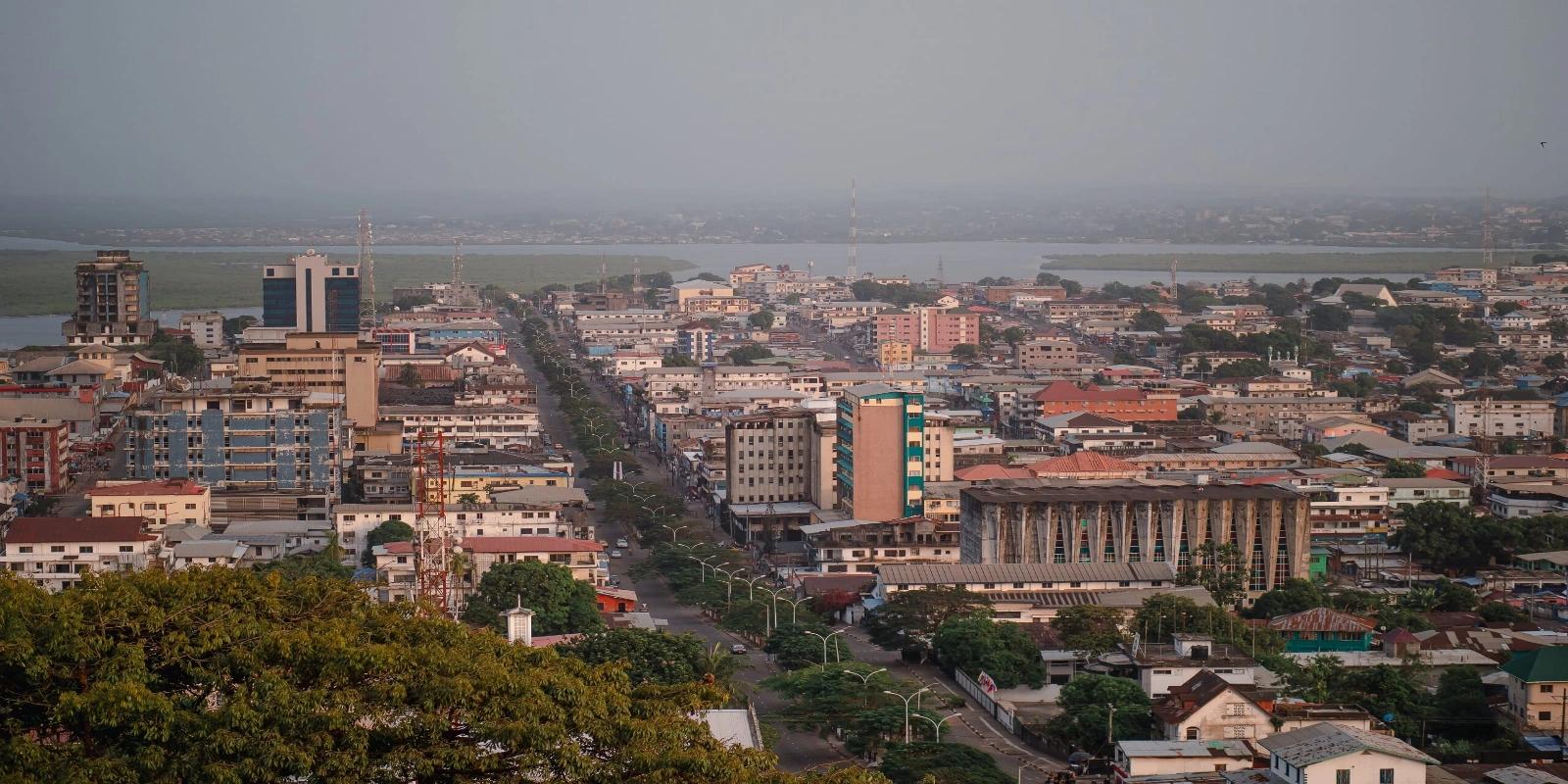 Panoramic view of the Monrovia, Liberia skyline with numerous buildings and a wide street leading towards a body of water under a hazy sky.