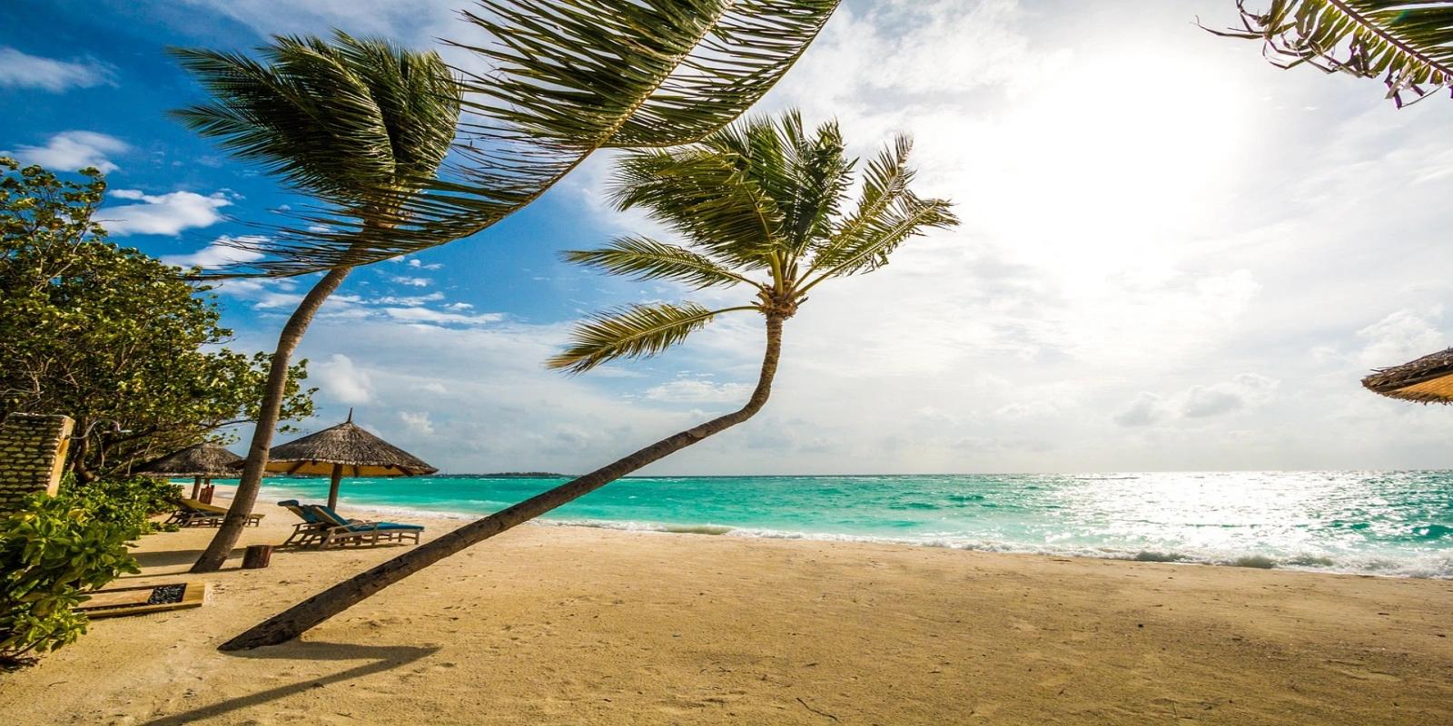 Tropical beach scene on Kuda Huraa Island, Maldives, with leaning palm trees, white sand, and turquoise ocean under a bright sky.