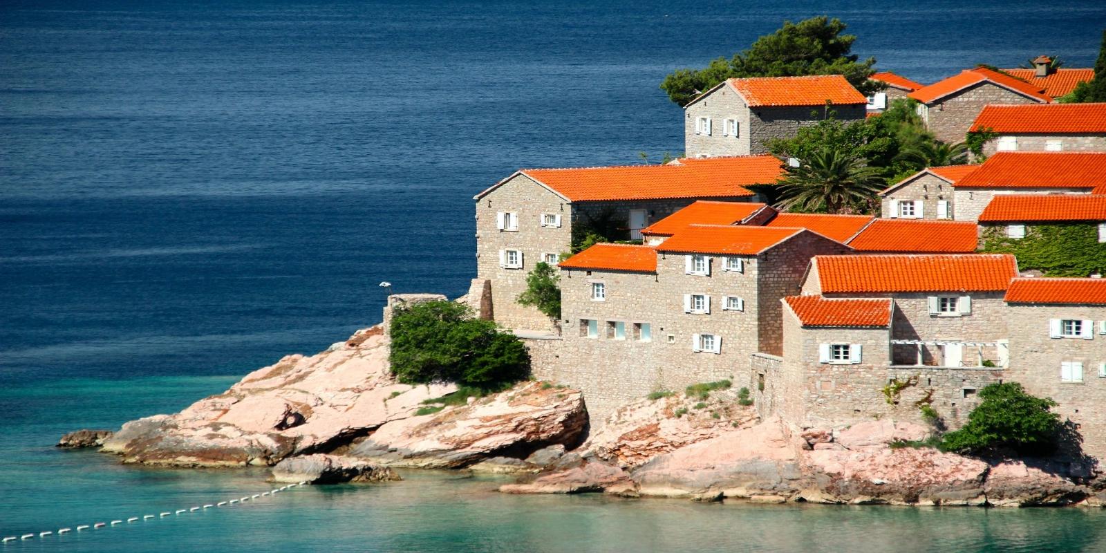 Aman Sveti Stefan, a picturesque fortified island village in Montenegro with traditional stone houses and orange-tiled roofs, nestled on a rocky outcrop by the clear blue sea.