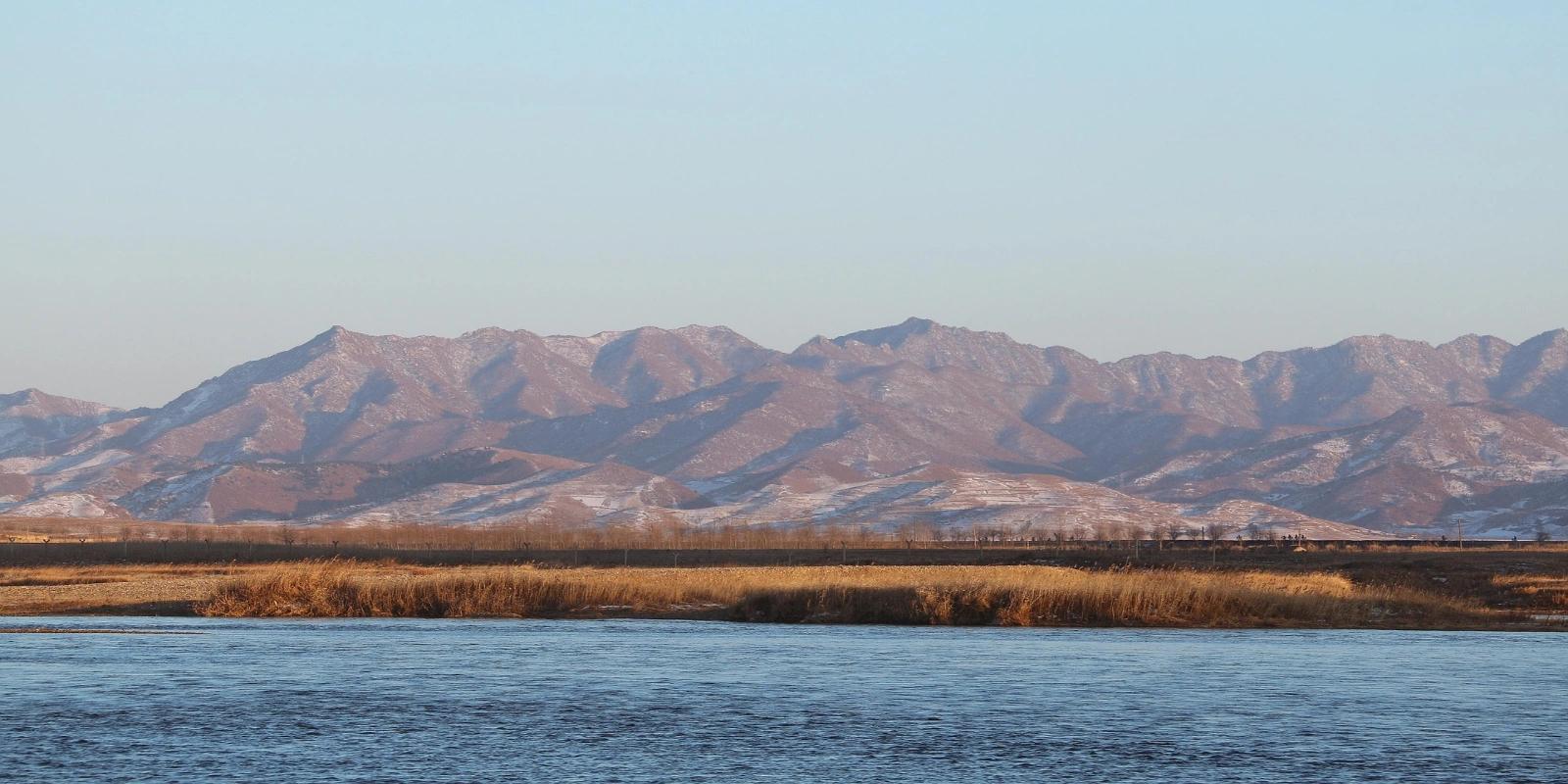 A wide river flows in the foreground with a dry, reedy bank, leading to snow-dusted mountains under a clear sky in North Korea.