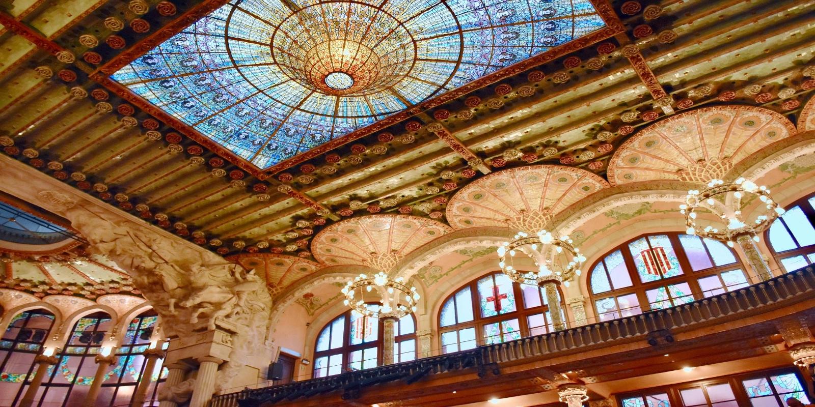 The ornate ceiling of the Palau de la Música Catalana in Palau, featuring a large stained-glass skylight and intricate architectural details with chandeliers.