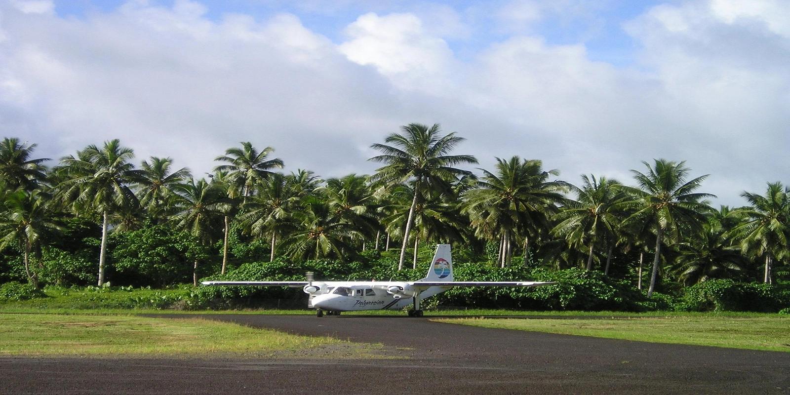 A small propeller plane parked on a runway in Samoa, surrounded by dense palm trees and lush green vegetation under a partly cloudy sky.