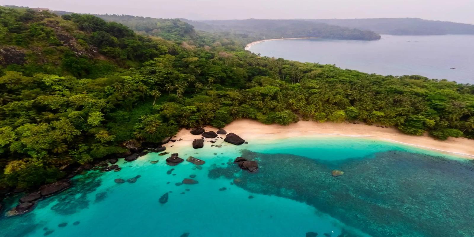 Aerial view of Praia das Bananas, a secluded cove on Principe Island in São Tomé and Príncipe, featuring a sandy beach, dark rocks, and clear turquoise waters, backed by a dense green forest.
