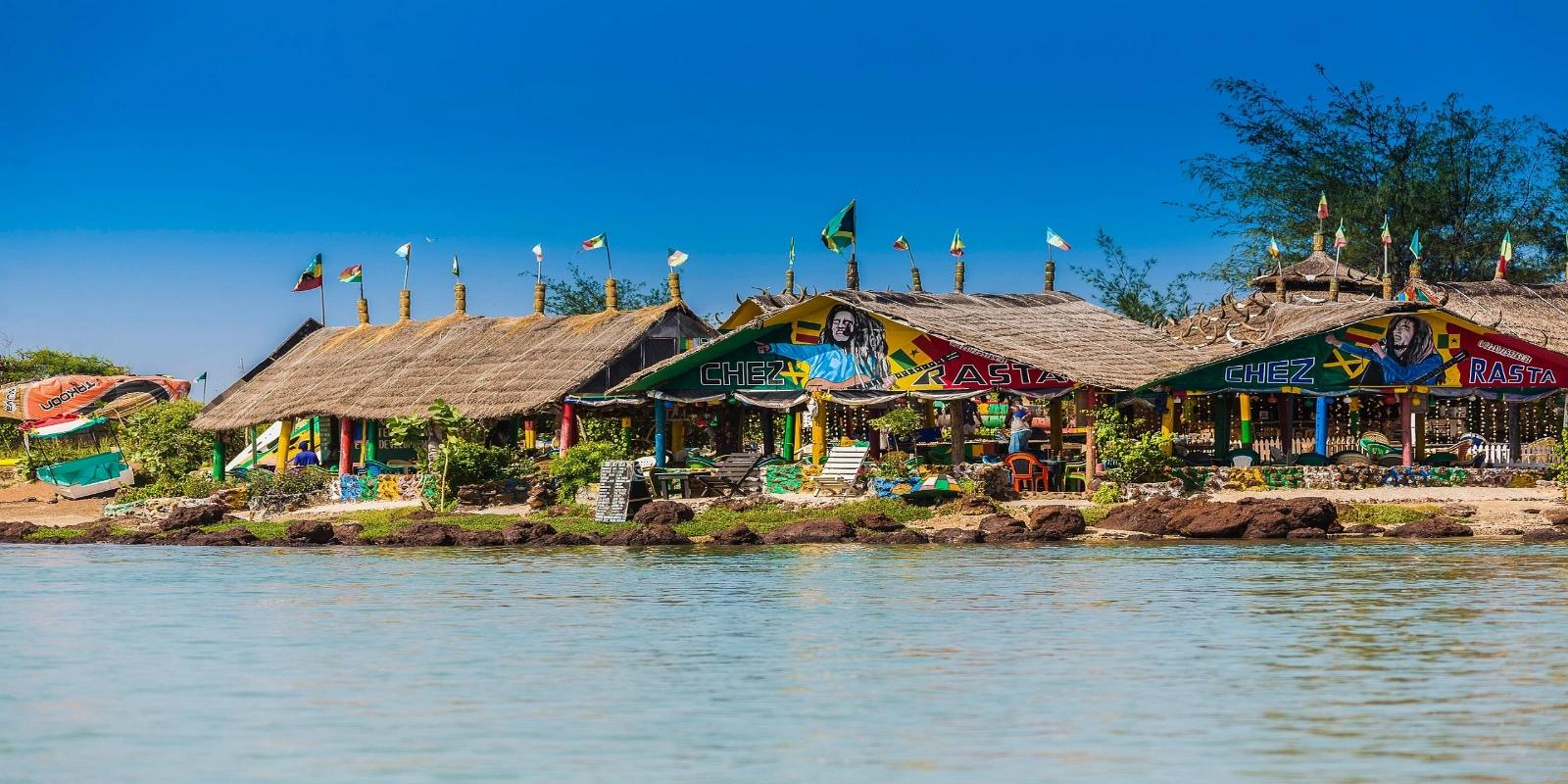 Colorful, thatched-roof buildings, a "Chez Rasta" themed establishment, lining the calm waterside in Senegal under a clear blue sky.