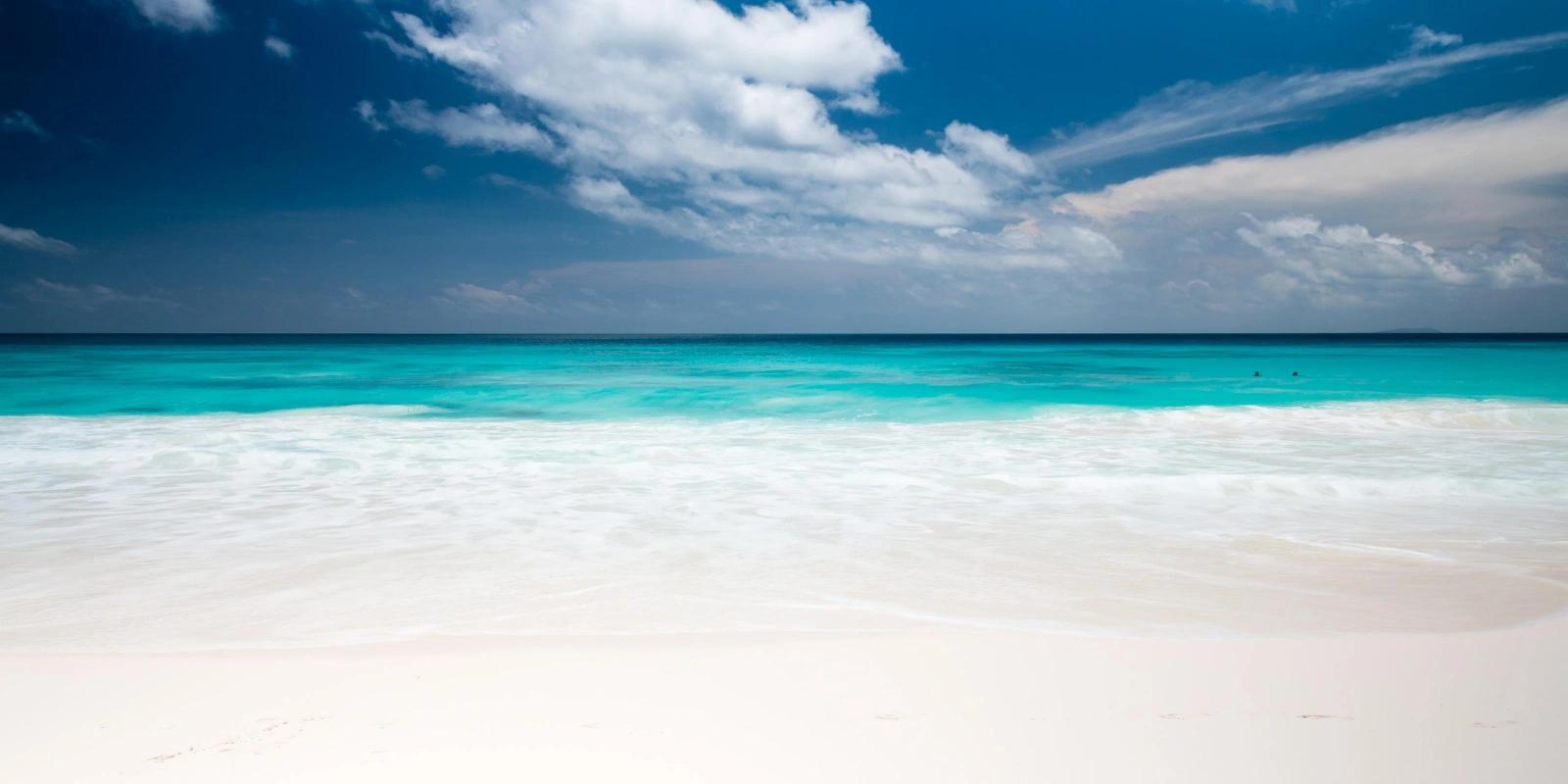 A pristine white sand beach meeting clear turquoise ocean water in the Seychelles, with gentle waves and a bright blue sky with white clouds.