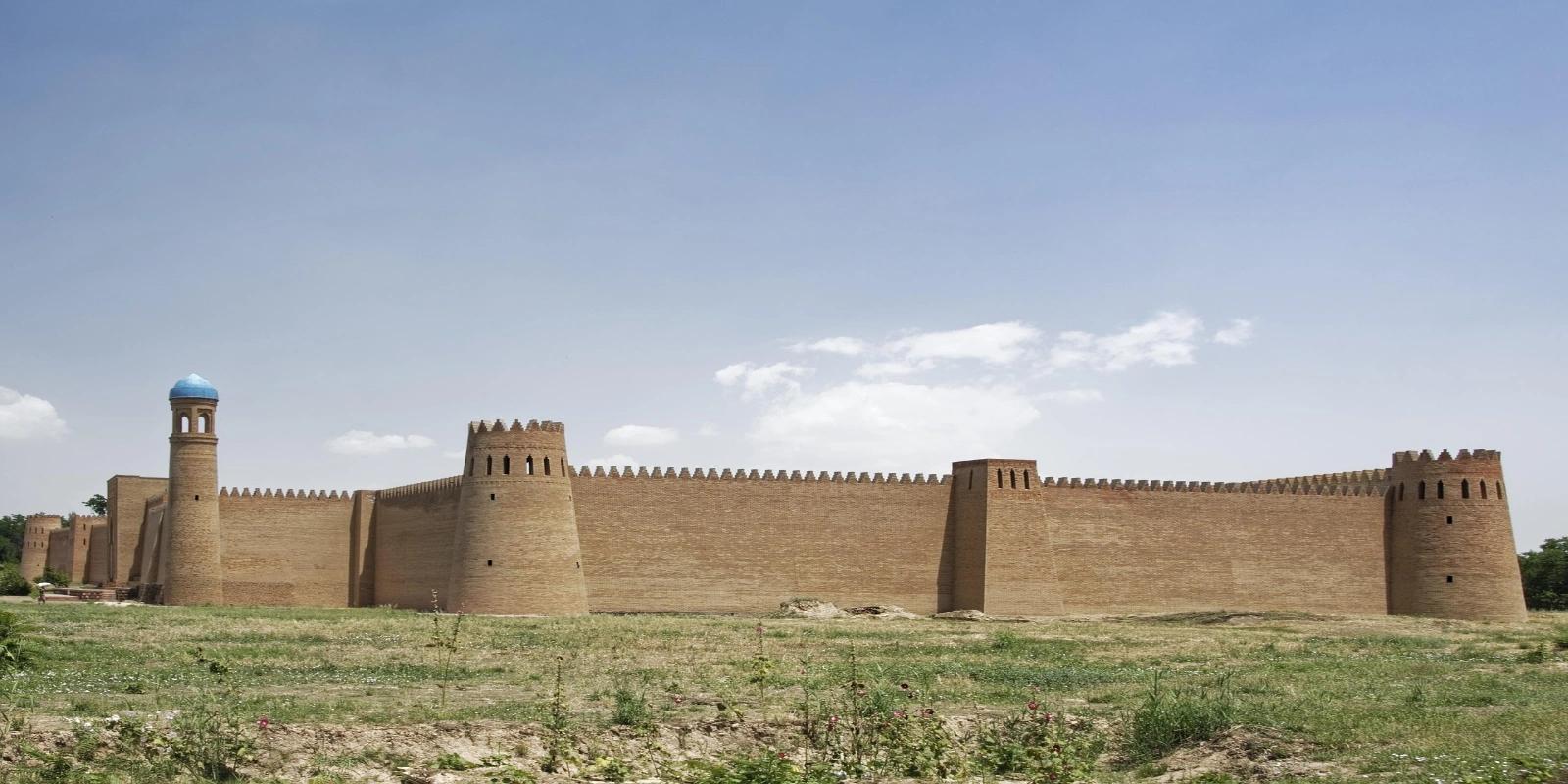 The imposing mud-brick walls and towers of Hulbuk Citadel in Tajikistan, standing on a grassy plain under a clear blue sky with sparse clouds.
