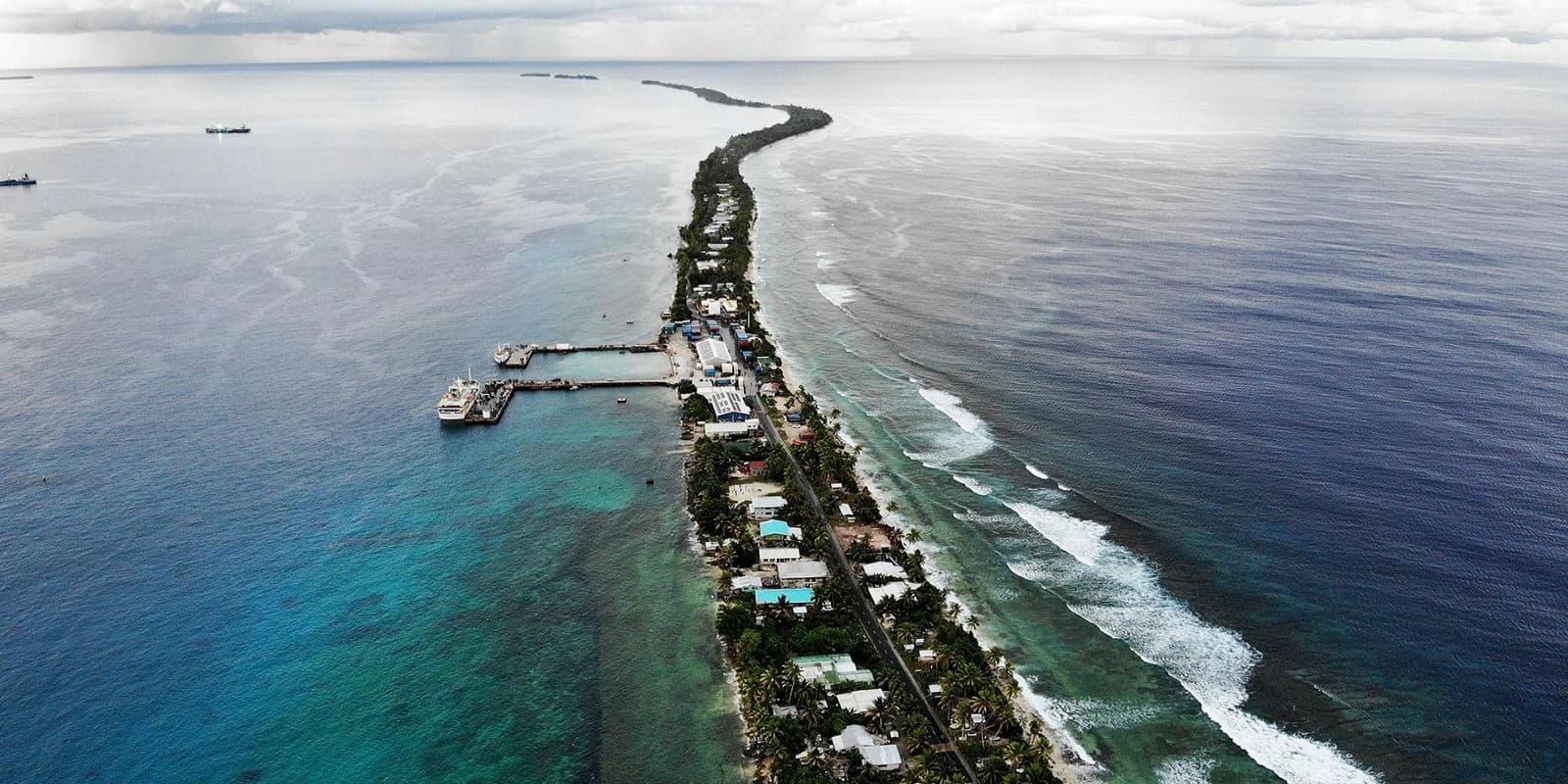 Aerial view of a narrow strip of land forming part of Tuvalu, with buildings and a road, surrounded by the vast blue Pacific Ocean with visible reef formations and waves.