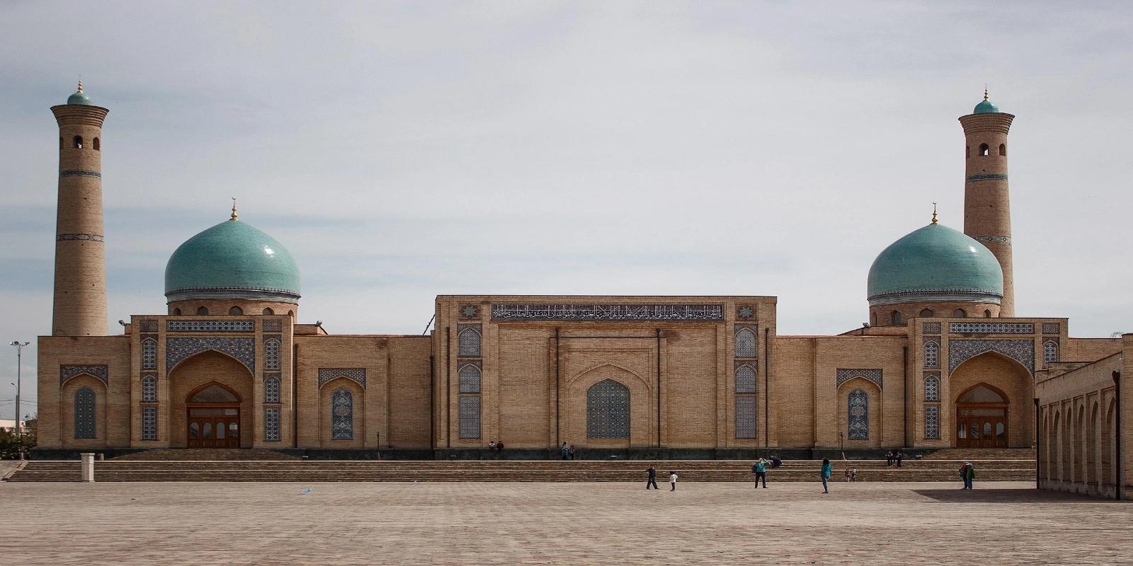 The grand architectural complex of the Tilla Sheikh Mosque in Tashkent, with two large turquoise domes, two tall minarets, and ornate brickwork, set against a light sky.
