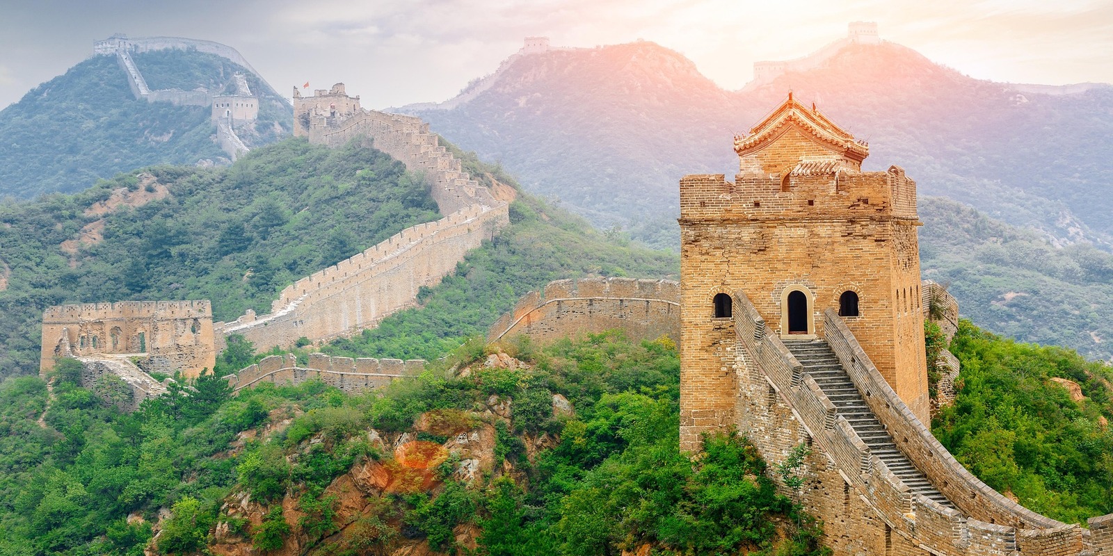 Great Wall of China winding across green mountain ridges with watchtower and sunset glow in background.
