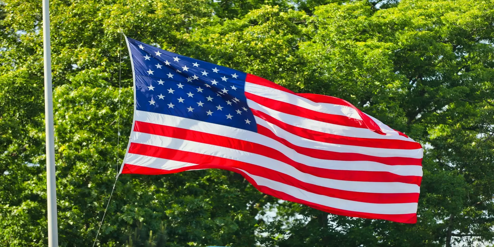 American flag waving in breeze against backdrop of green trees on sunny day.
