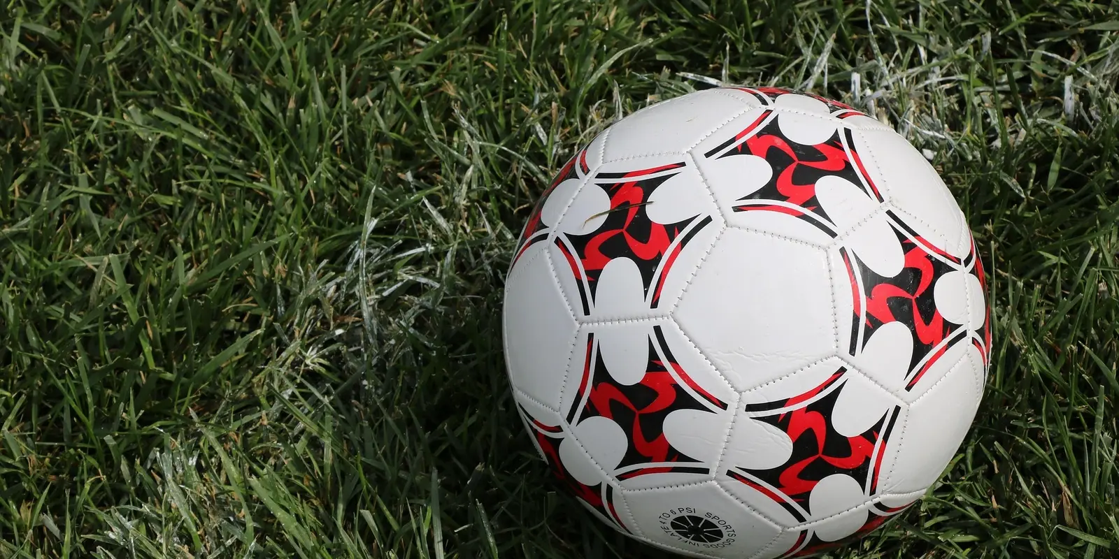 A close-up of a soccer ball with a red, black, and white pattern resting on a green grass field near a white boundary line.