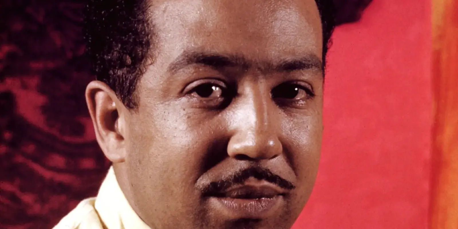 Close-up portrait of Langston Hughes with serious expression against red background.
