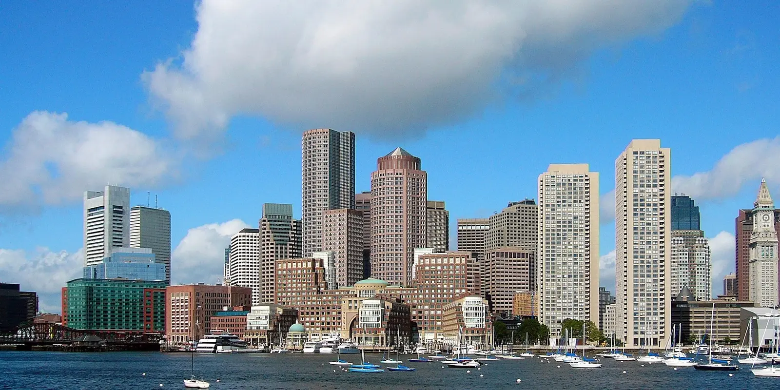 Boston skyline view from harbor with sailboats in foreground and downtown skyscrapers under blue sky.