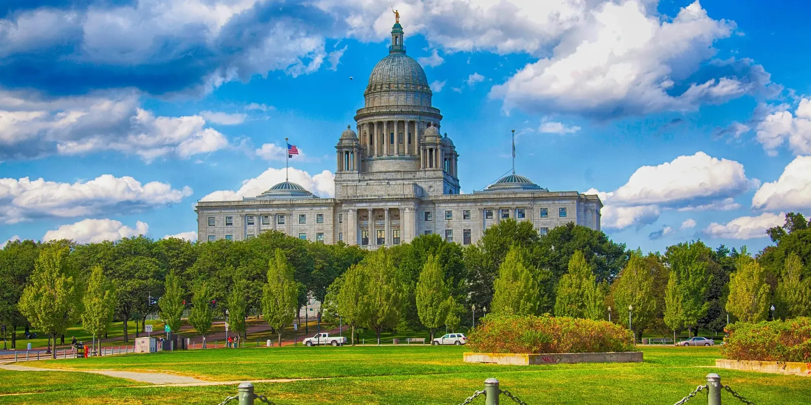 A vibrant view of the Rhode Island State House, a grand white-domed building, surrounded by lush green trees and a manicured lawn under a partly cloudy sky.