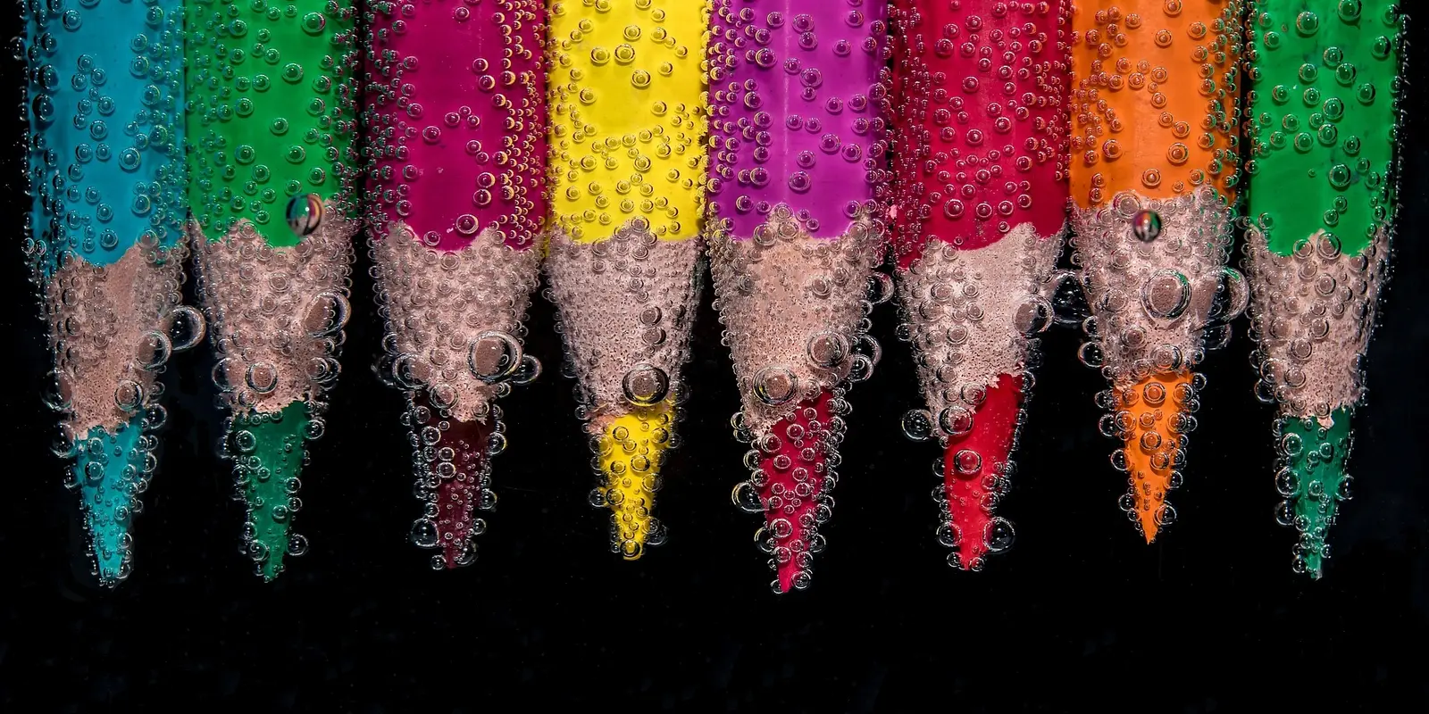 A close-up photograph of sharpened colored pencils submerged in water, covered in tiny air bubbles. The pencils display vibrant colors, including blue, green, yellow, pink, red, and orange, against a dark background, creating a striking contrast.