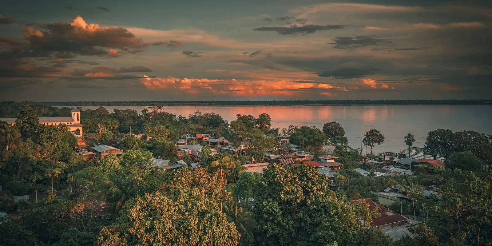 A scenic view of an Amazonian village in Colombia at sunset, with lush green trees, rustic houses, and a calm river reflecting the golden hues of the sky.