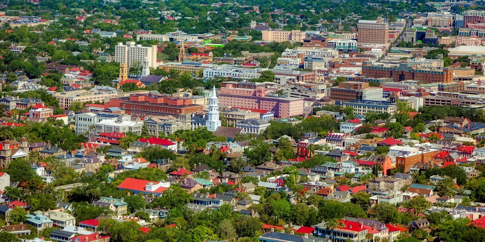 A colorful aerial view of Charleston, South Carolina, showcasing historic buildings, lush greenery, and a mix of red, blue, and pastel-colored rooftops.