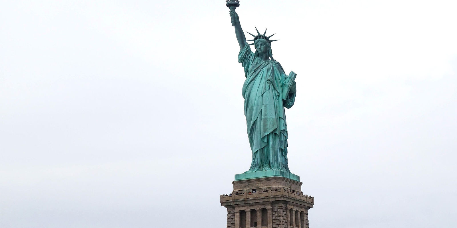 The Statue of Liberty standing tall on Liberty Island, New York, with a cloudy sky in the background. The green patina of the statue contrasts with the golden flame of the torch.