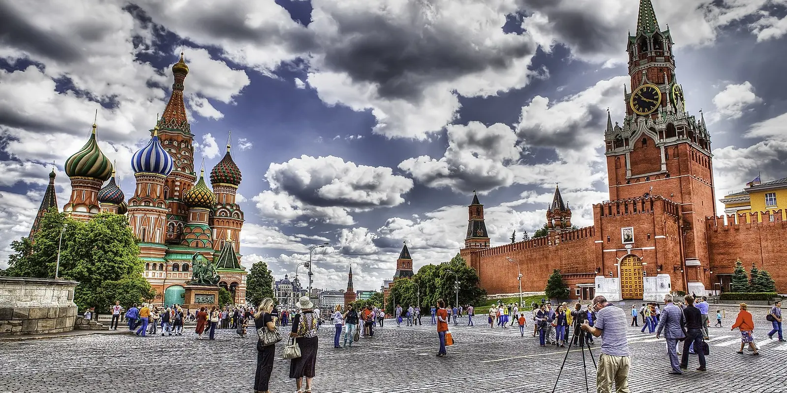 A vibrant view of Red Square in Moscow, Russia, featuring the colorful domes of St. Basil's Cathedral on the left and the Spasskaya Tower of the Kremlin on the right, with many tourists strolling across the cobblestone square under a dramatic sky with clouds.