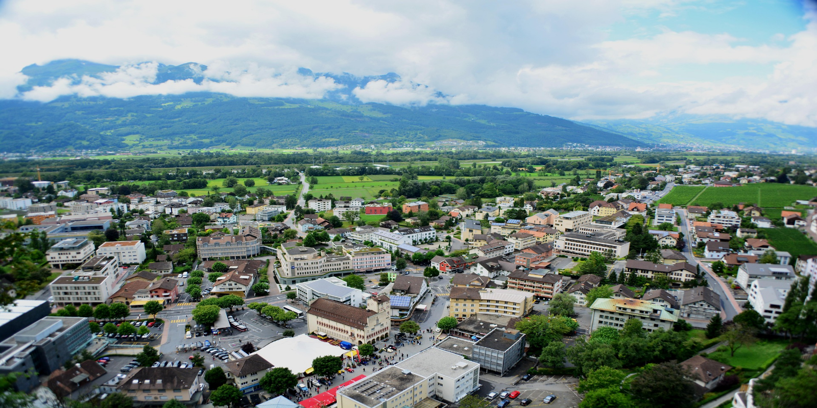 Panoramic view of Vaduz, Liechtenstein, showing a town with various buildings and surrounding green fields nestled at the foot of mountains under a cloudy sky.